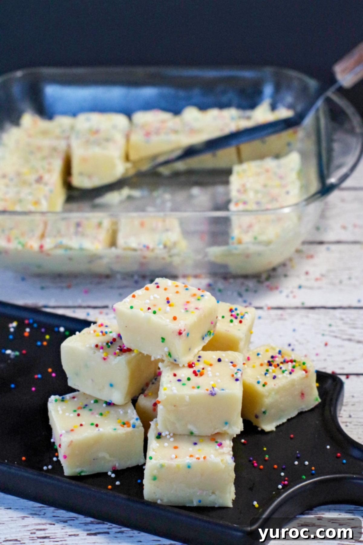 Easy Sugar Cookie Fudge 8 A beautiful arrangement of sugar cookie fudge squares on a black platter in the foreground, with a larger square glass container of fudge softly blurred in the background, ready for enjoyment.