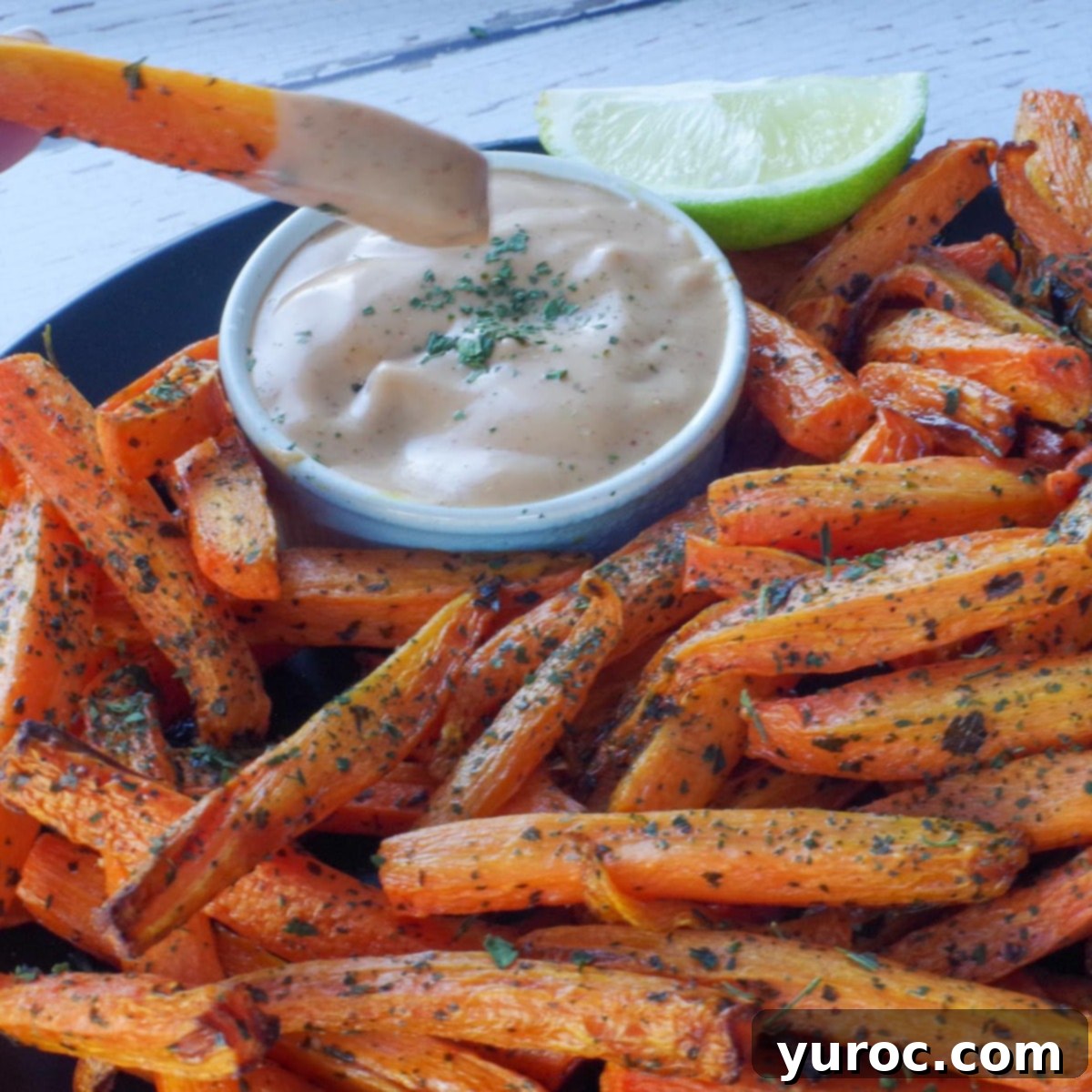 A carrot fry being dipped into a creamy dip, with a plate of more carrot fries in the background.
