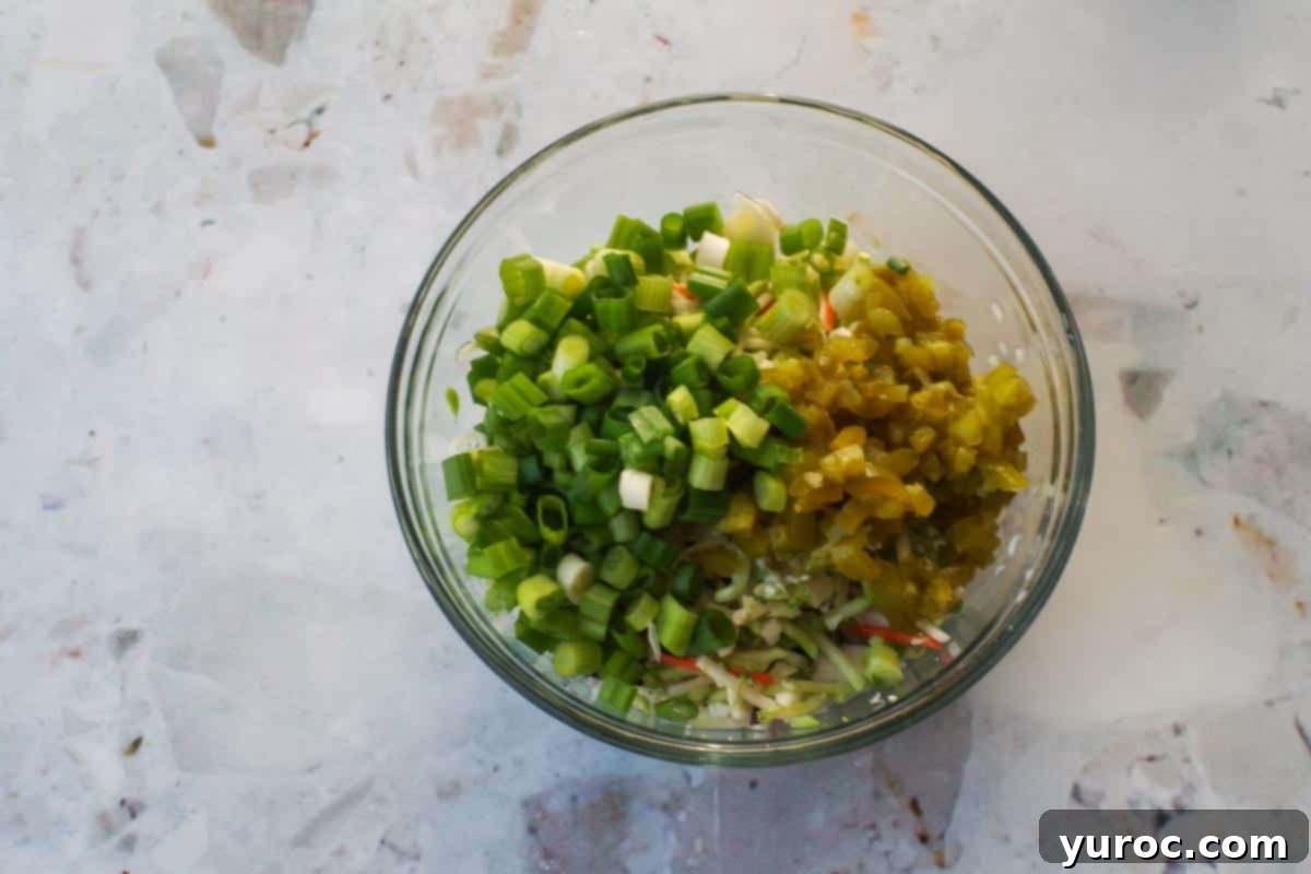 Chopped green onions, pepperoncini, and tamed jalapenos added to the glass bowl with broccoli slaw.