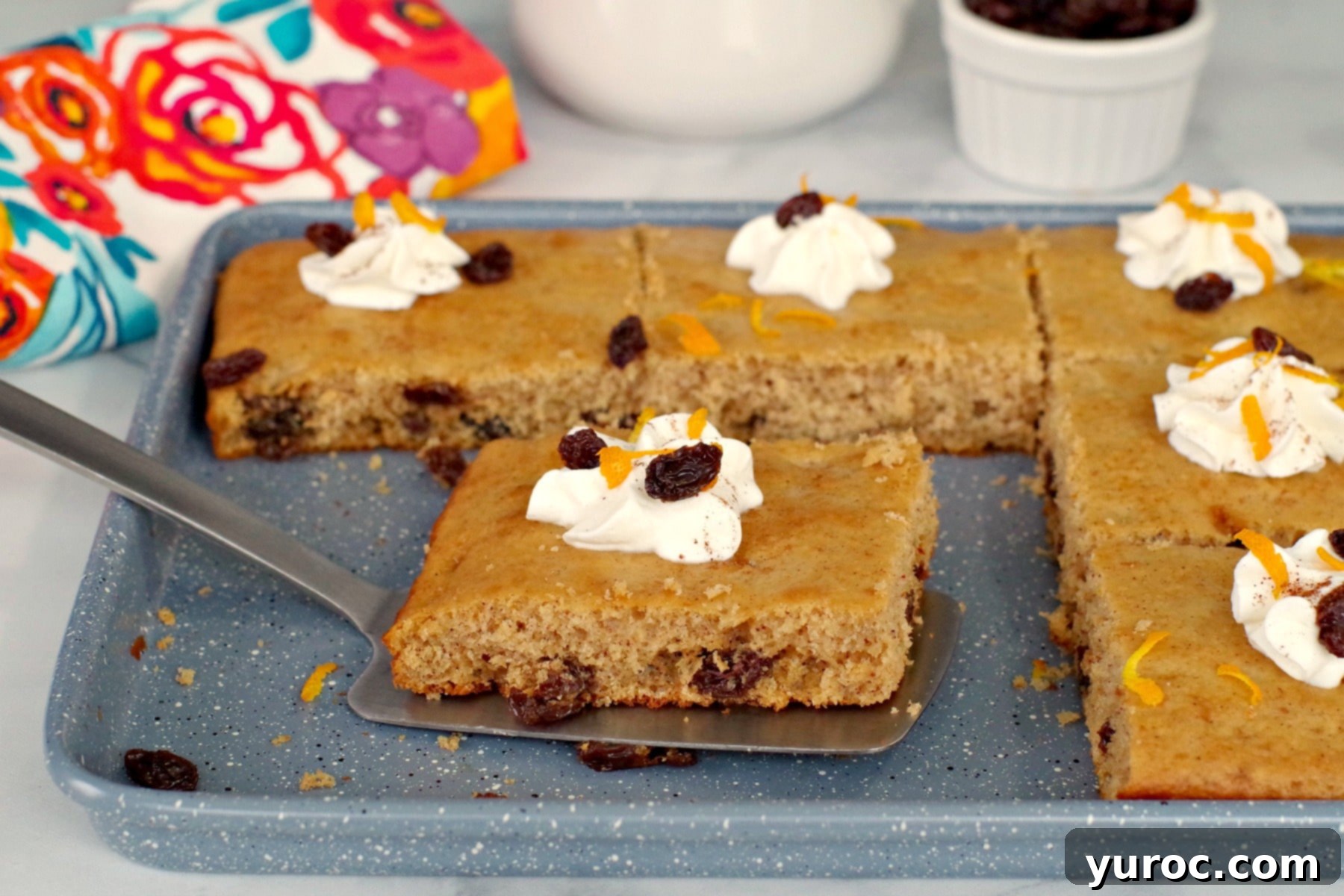 A golden brown Rum & Raisin sheet pan pancake piece, perfectly baked, being lifted off a blue baking sheet with a spatula, ready to be served.