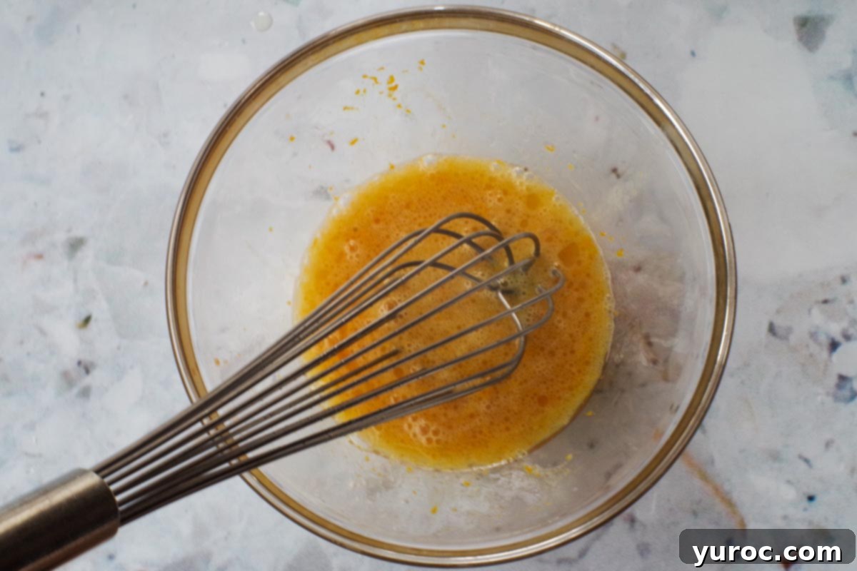Wet ingredients, including eggs, oil, vanilla, rum extracts, and orange zest, perfectly whisked together in a medium glass bowl.