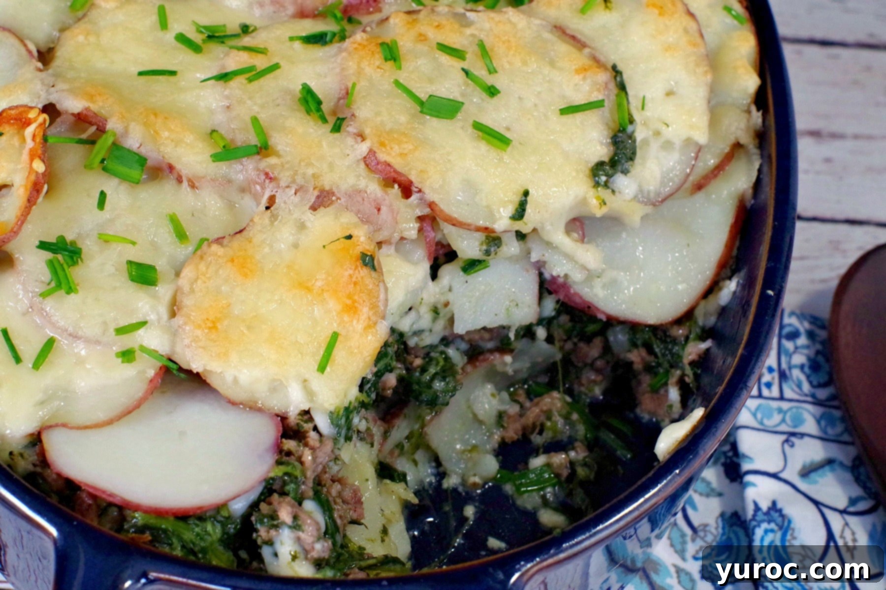 A serving of the delicious Potato and Ground Beef Casserole with Spinach on a white plate, showing golden-brown cheese, tender potatoes, and savory beef, with a portion already scooped from the main blue casserole dish.
