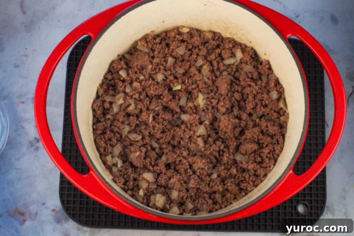 Homestyle Hamburger Potato Soup 5 Ground beef and chopped onion cooking together in a vibrant orange Dutch oven on the stovetop.