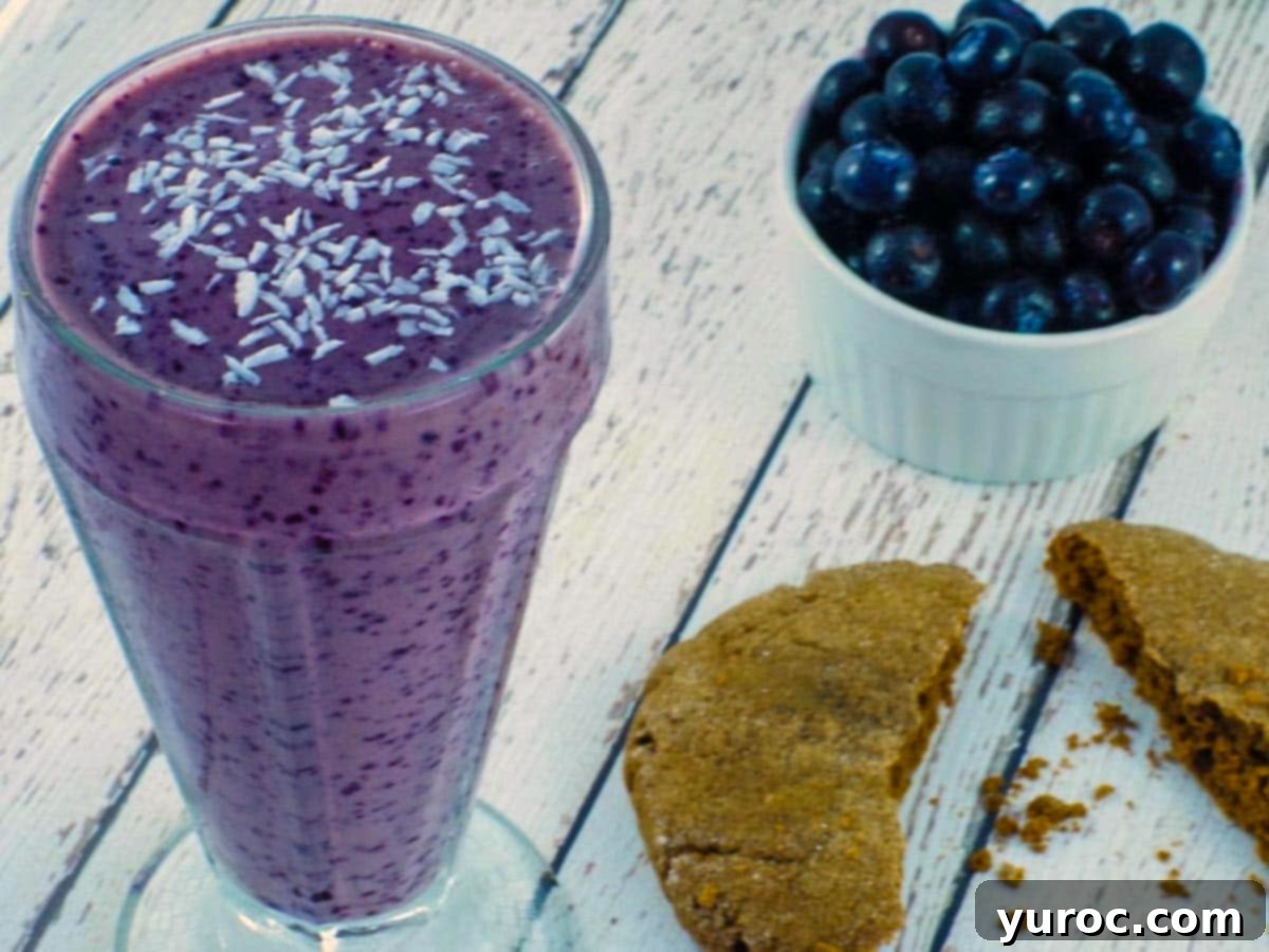 Vibrant blueberry smoothie with almond milk, garnished with shredded coconut, set on a rustic white wooden surface. Fresh blueberries and a half-eaten cookie are visible in the soft-focus background, enhancing the smoothie's delicious appeal.