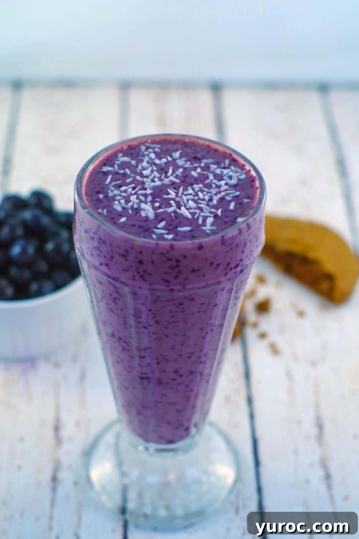 A close-up shot of a creamy blueberry smoothie with almond milk, topped with shredded coconut. The smoothie is in a clear glass on a white faux wooden surface, with a half a cookie and fresh blueberries in the background. This image highlights the smoothie's inviting texture and garnish.