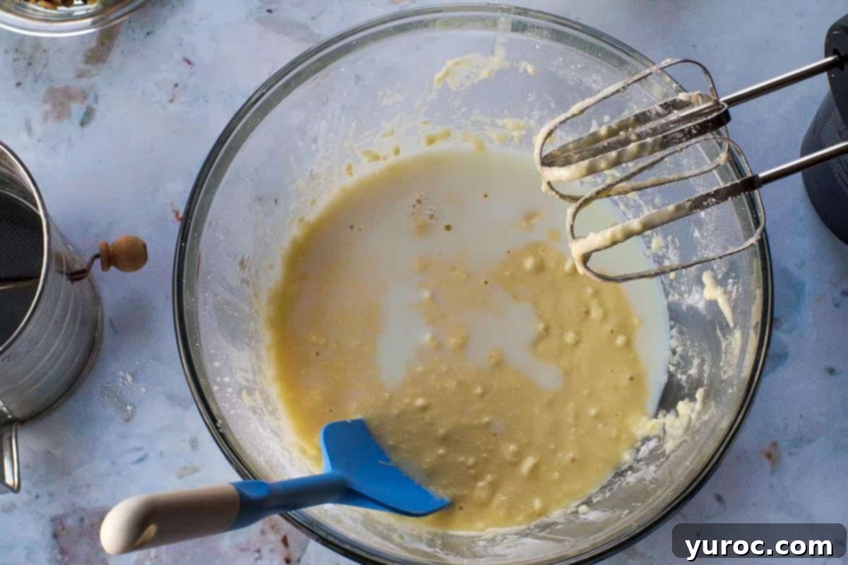 Milk being added to the wet mixture in a large glass bowl during alternate additions with flour