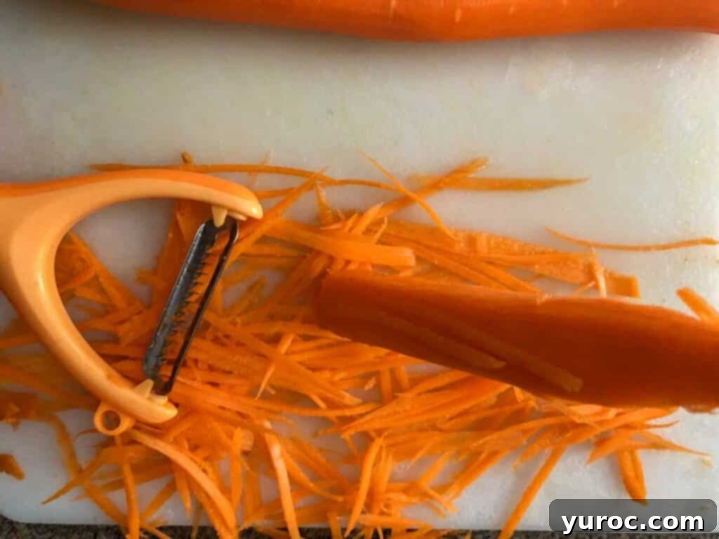 A carrot being shredded into thin strips using a julienne peeler on a white cutting board