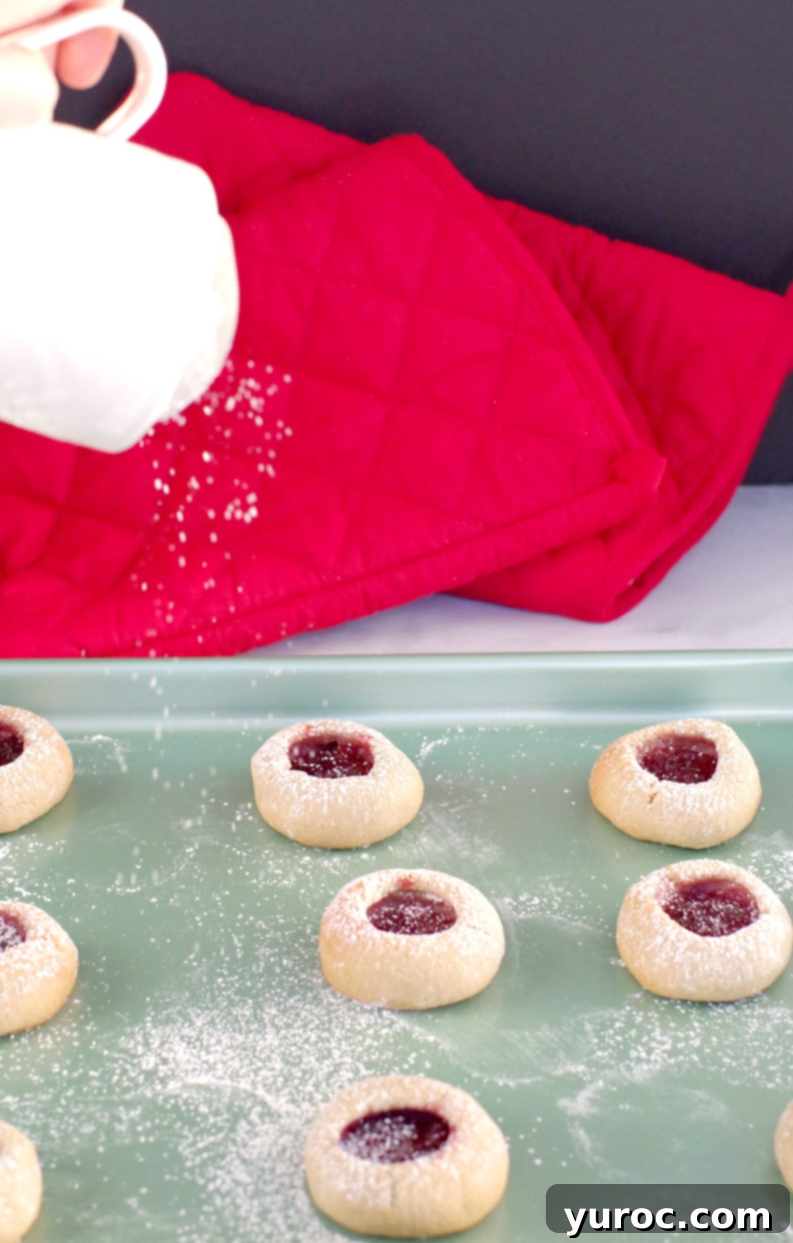 Freshly baked jam cookies cooling on a green cookie sheet, with a delicate dusting of powdered sugar being applied.
