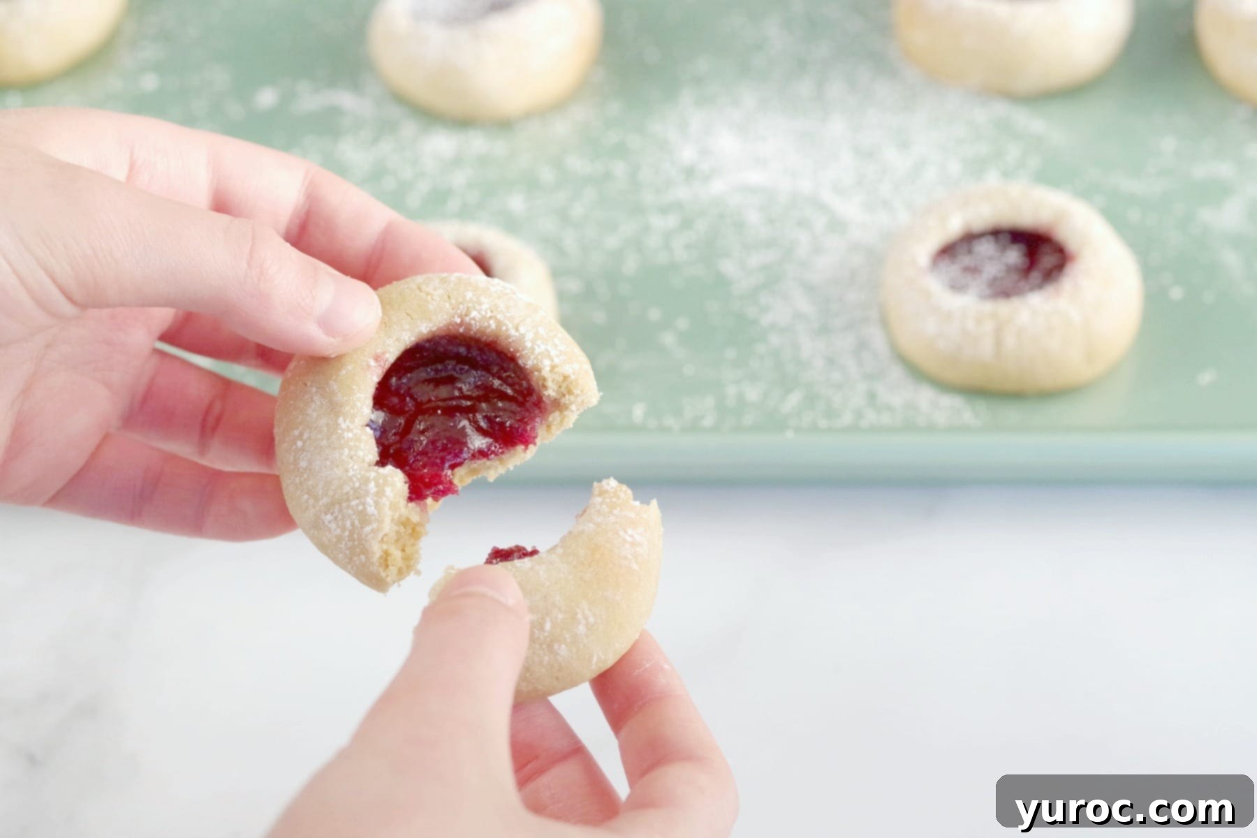 A raspberry jam cookie being gently broken in half, showcasing its soft interior and vibrant jam filling.