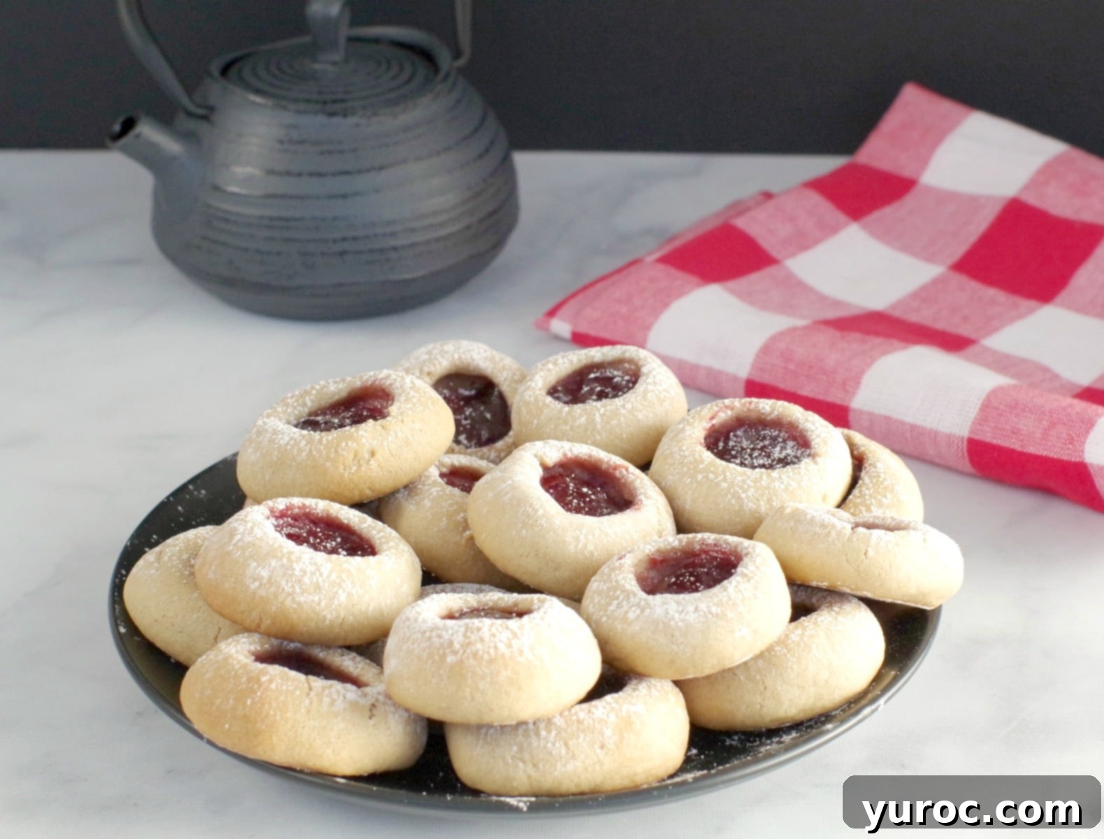 A black plate filled with golden-brown raspberry jam thumbprint cookies, alongside a dark teapot and a red checkered tea towel, suggesting a cozy afternoon treat.