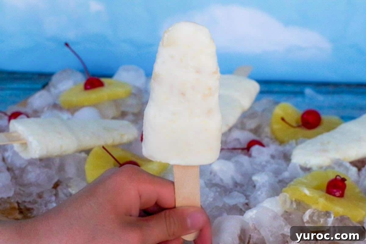 Cool & Creamy Virgin Pina Colada Pops 4 Hand holding a creamy Piña Colada popsicle against a backdrop of fresh pineapple, cherries, and more popsicles on ice.
