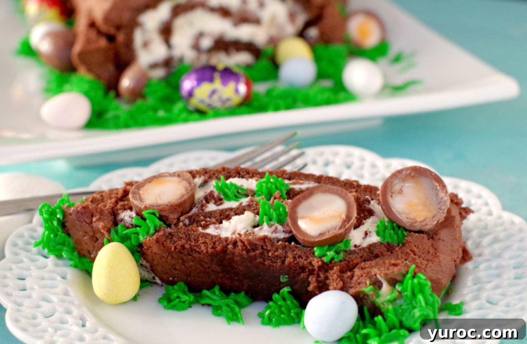 Delicious Easter Creme Egg Swiss Roll Cake on a white plate with a slice cut, showcasing the beautiful swirl and the whole cake in the background.