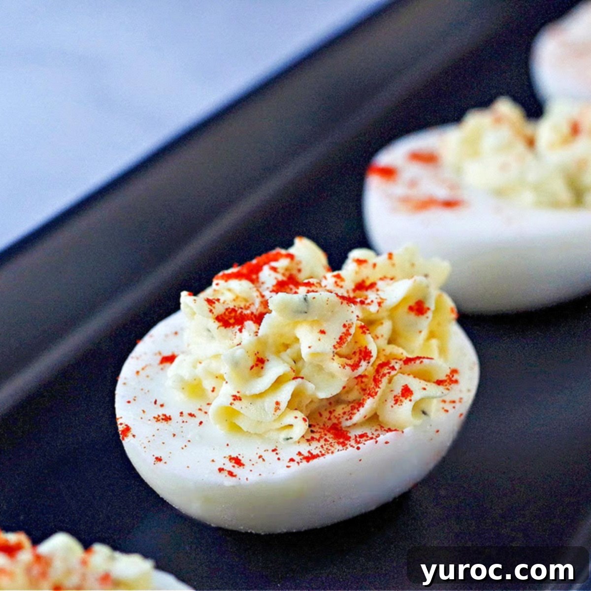 Close-up of a deviled egg with paprika sprinkled on top, resting on a black serving tray, with other deviled eggs blurred in the background.