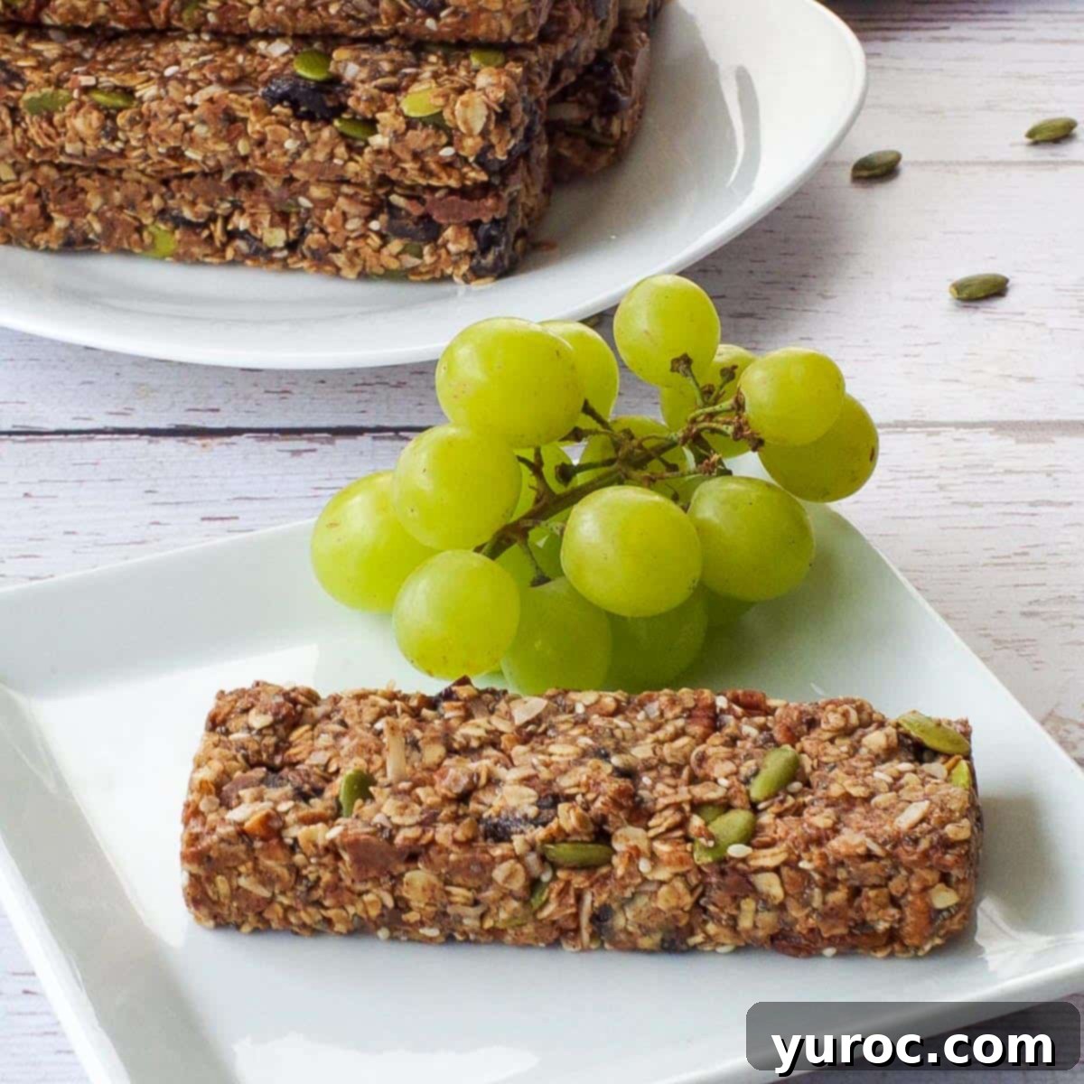 Ultimate Turkey Bacon Ranch Wrap 9 A single almond butter granola bar on a white plate with green grapes, and a pile of granola bars in the background.