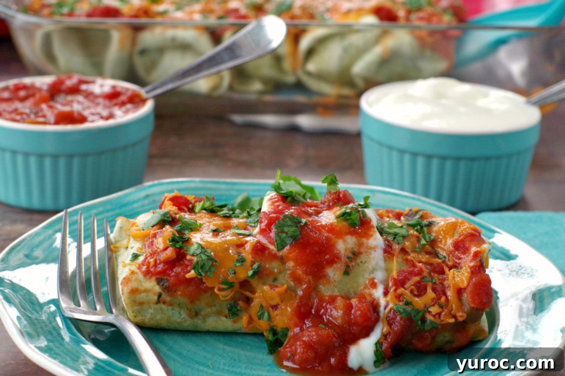 A single serving of healthy leftover turkey enchiladas on a blue plate, with a glass baking dish of enchiladas, sour cream, and salsa in blue ramekins in the background, showcasing a delicious and satisfying meal.