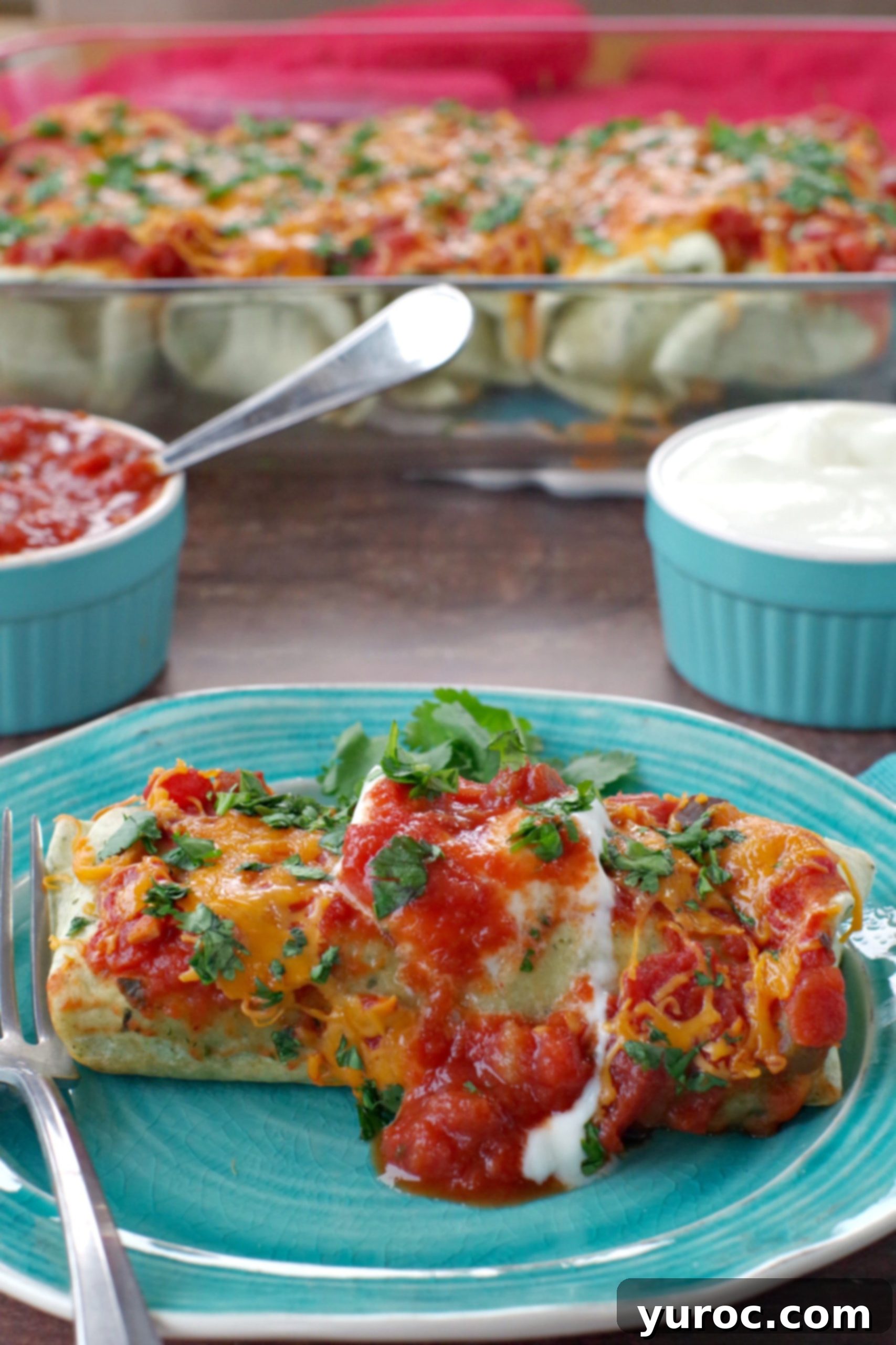 A single delicious and healthy leftover turkey enchilada on a blue plate, with a full glass baking dish of enchiladas and blue ramekins holding sour cream and salsa in the background, perfect for a Weight Watchers friendly meal.