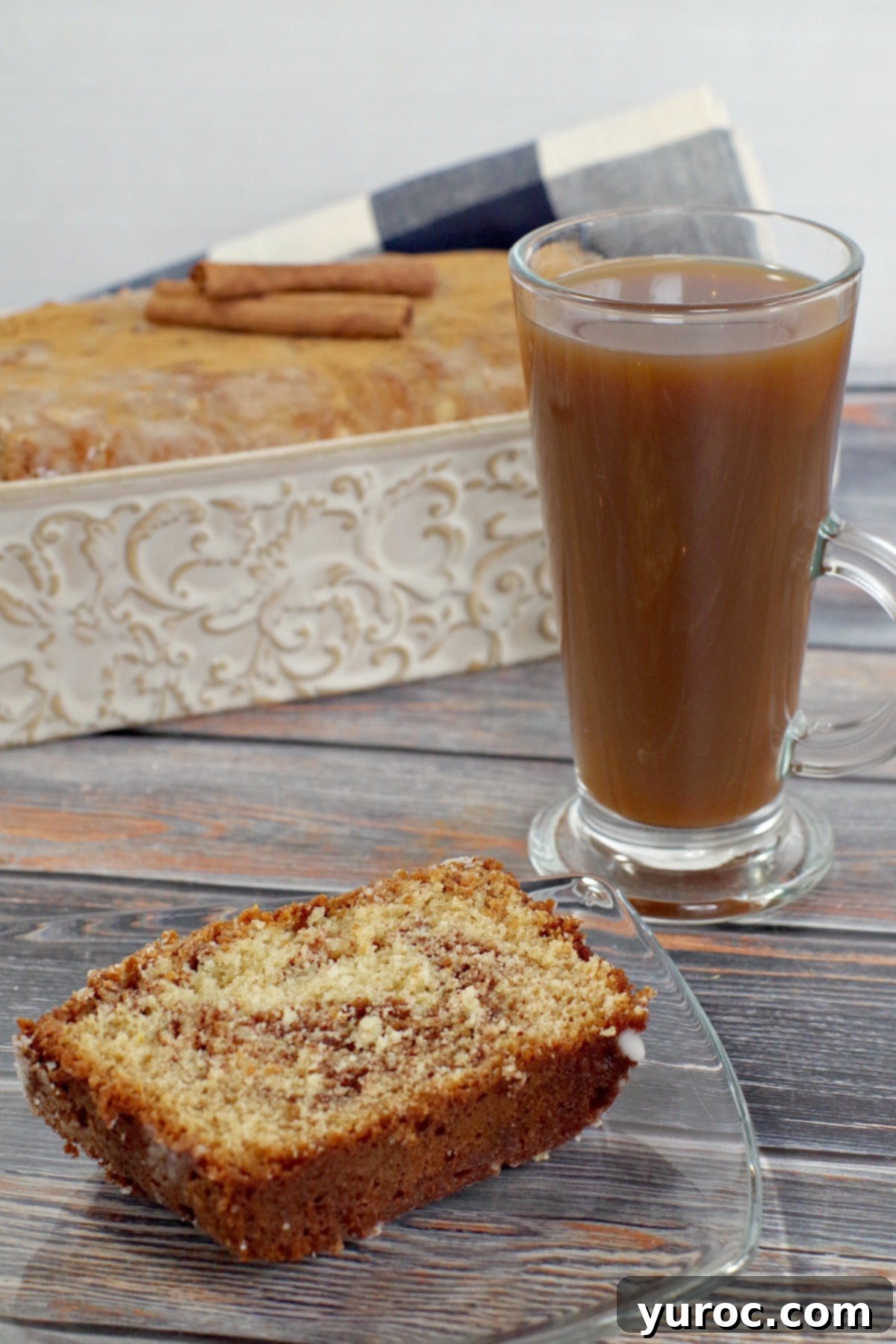 slice of cinnamon bread on a glass plate next to a cup of coffee and loaf pan in background