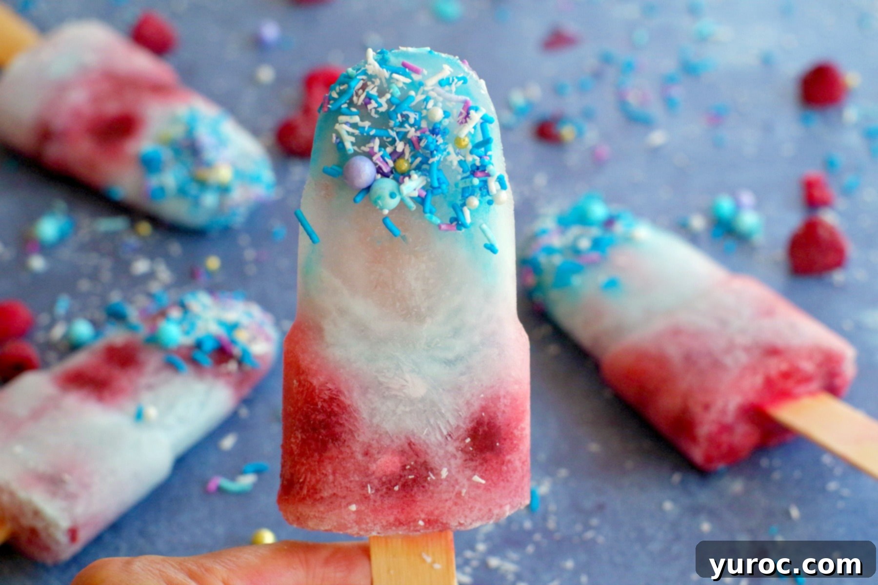 A refreshing Coconut Blue Raspberry Popsicle held against a blurred background of other popsicles, ready to be enjoyed.