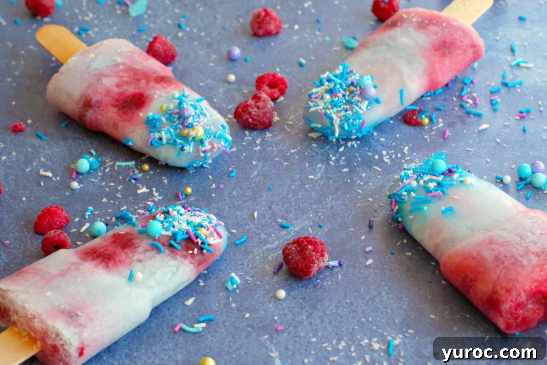 Four vibrant blue raspberry popsicles arranged on a blue surface, surrounded by fresh coconut, frozen raspberries, and blue sprinkles.