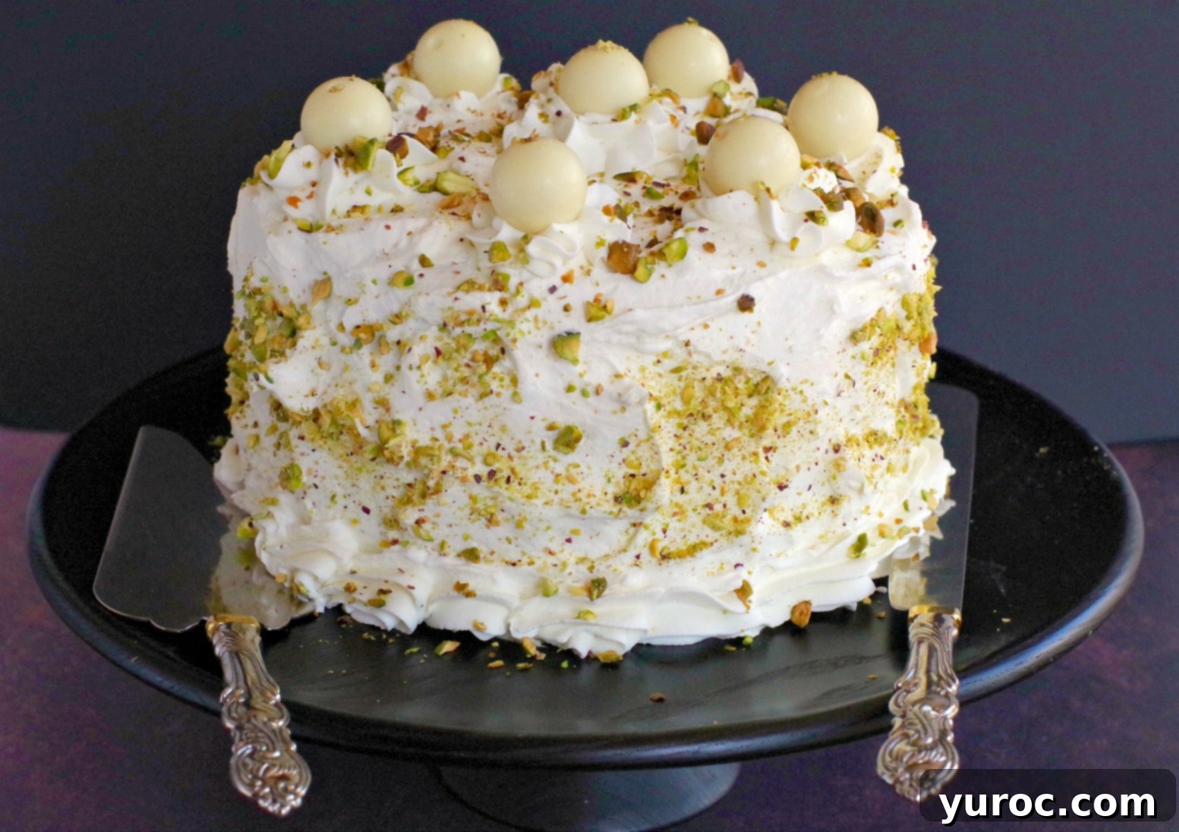 A stunning three-layer White Chocolate Pistachio Cake, beautifully frosted and garnished, resting on a black cake stand with a cake lifter and knife nearby. Perfect for celebrations and special events.