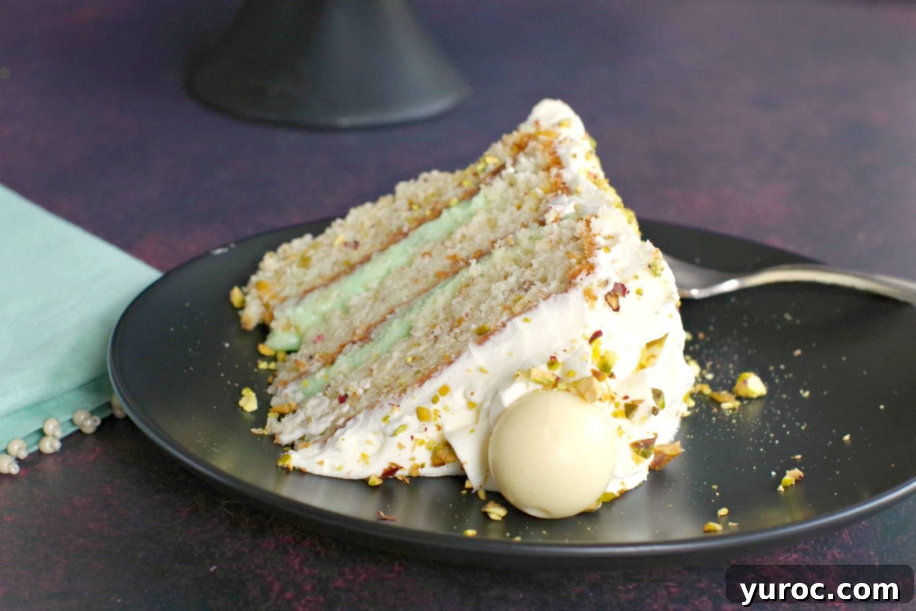 A close-up shot of a perfect slice of White Chocolate Pistachio Cake, revealing the moist cake layers, vibrant green pistachio pudding filling, and creamy white chocolate frosting on a black plate.
