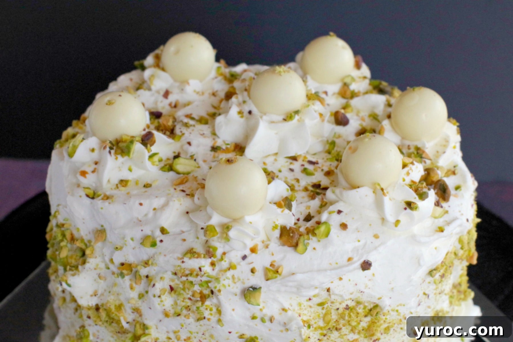 The impressive top view of the White Chocolate Pistachio Cake, showcasing its smooth white chocolate frosting, delicate piped dollops, and a generous sprinkle of chopped pistachios on a black cake stand, ready for slicing.