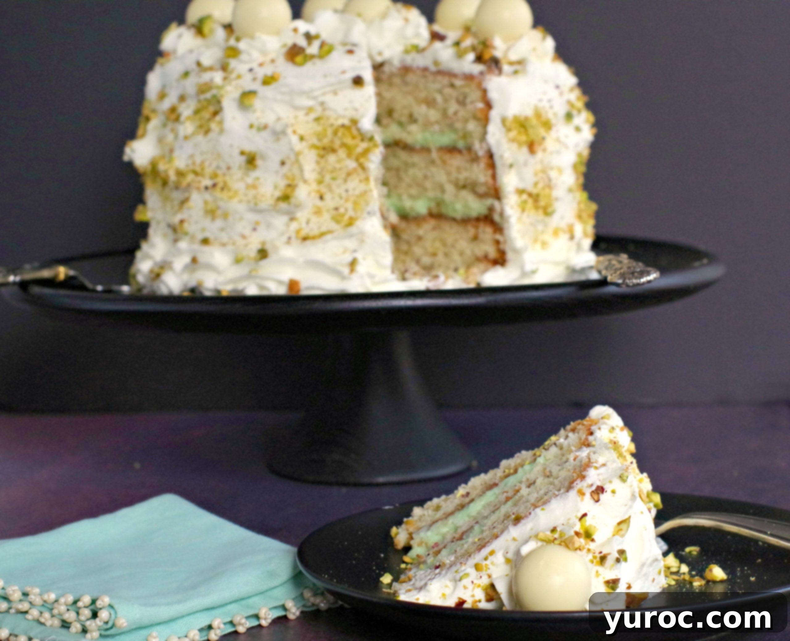 A beautifully arranged scene with the full White Chocolate Pistachio Cake on a black tray in the background, and a single, perfectly cut slice in the foreground, showcasing its inviting layers and textures.