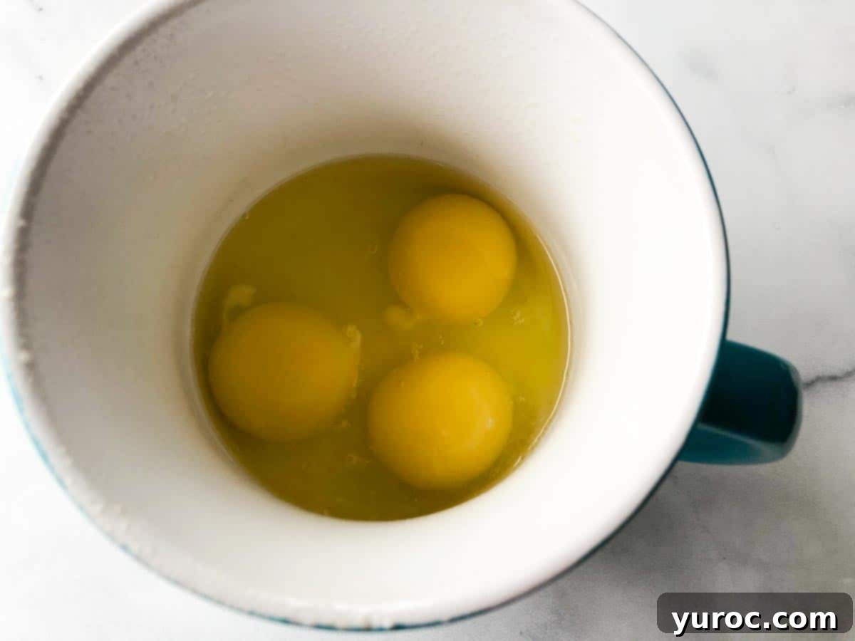 Three fresh eggs cracked into a large, microwave-safe mug, prepared with non-stick spray.