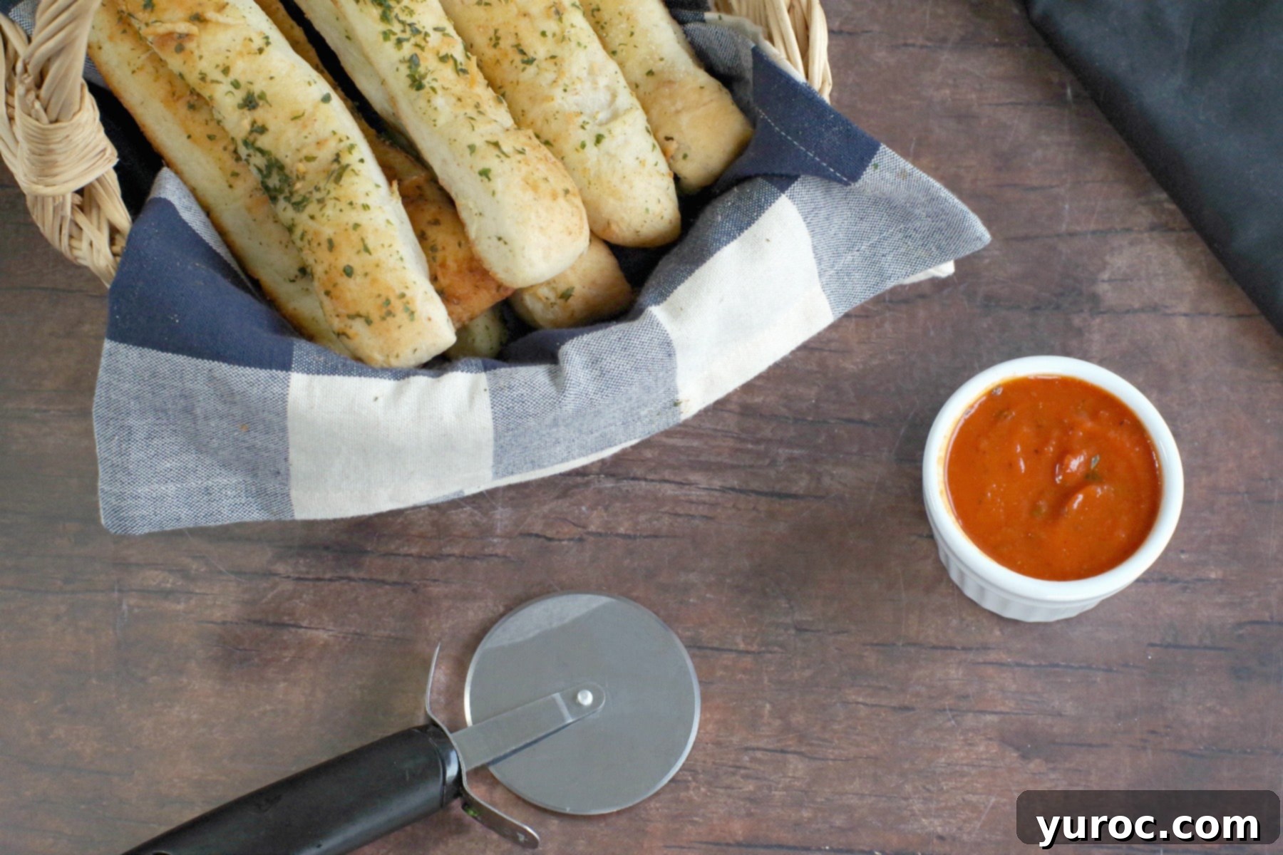 Italian breadsticks in a rustic basket, accompanied by a small bowl of savory pizza sauce and a pizza cutter on the side.