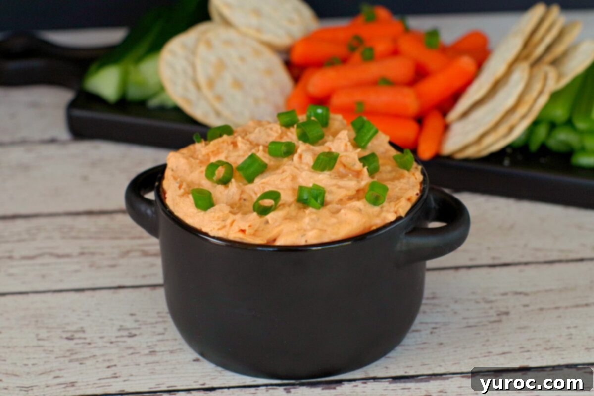 Creamy buffalo chicken dip served in a black bowl, garnished with green onions, with fresh vegetables and crackers in the background.