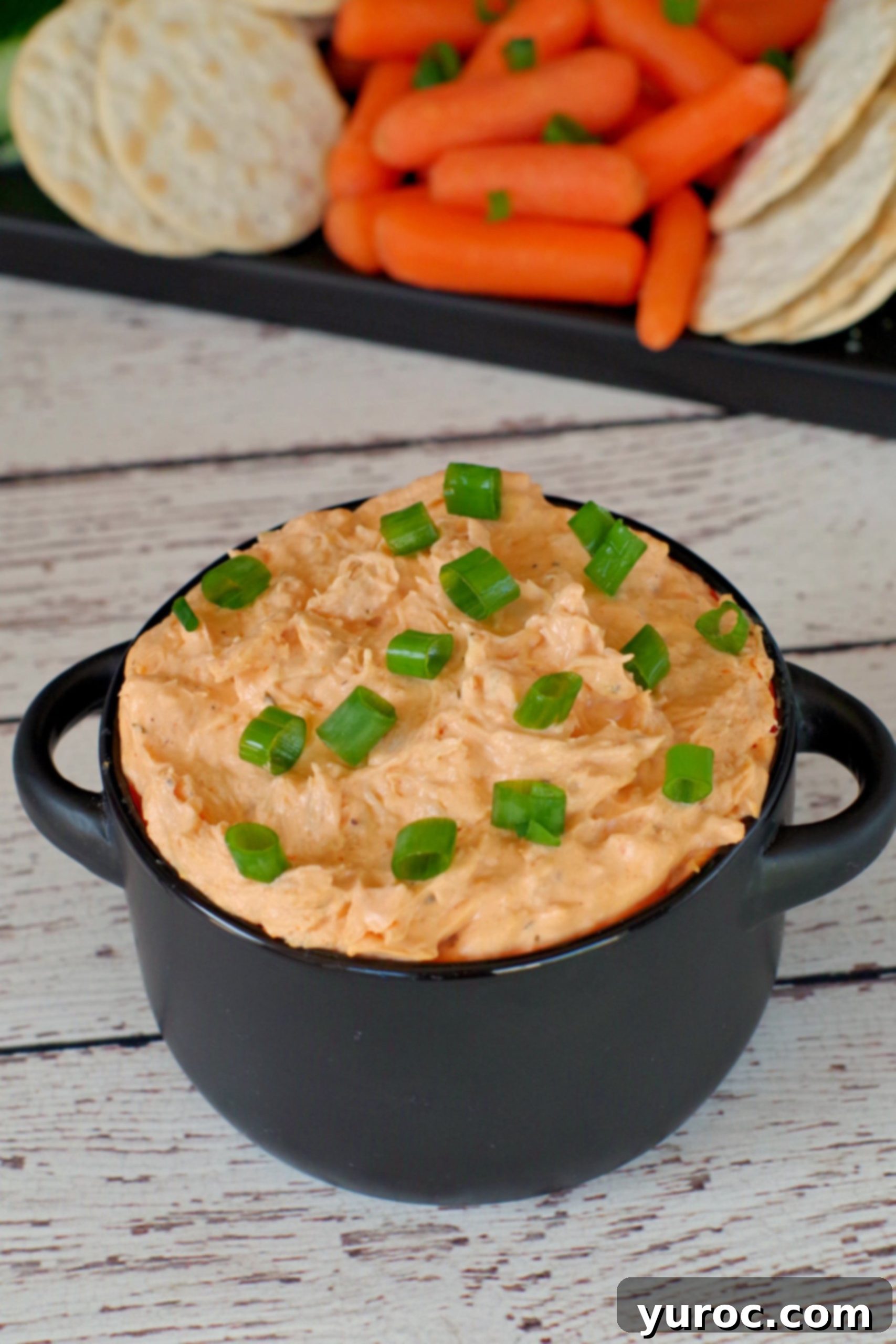 A black serving bowl filled with creamy buffalo chicken dip, surrounded by an assortment of fresh vegetables like celery and carrots, and various crackers for dipping.
