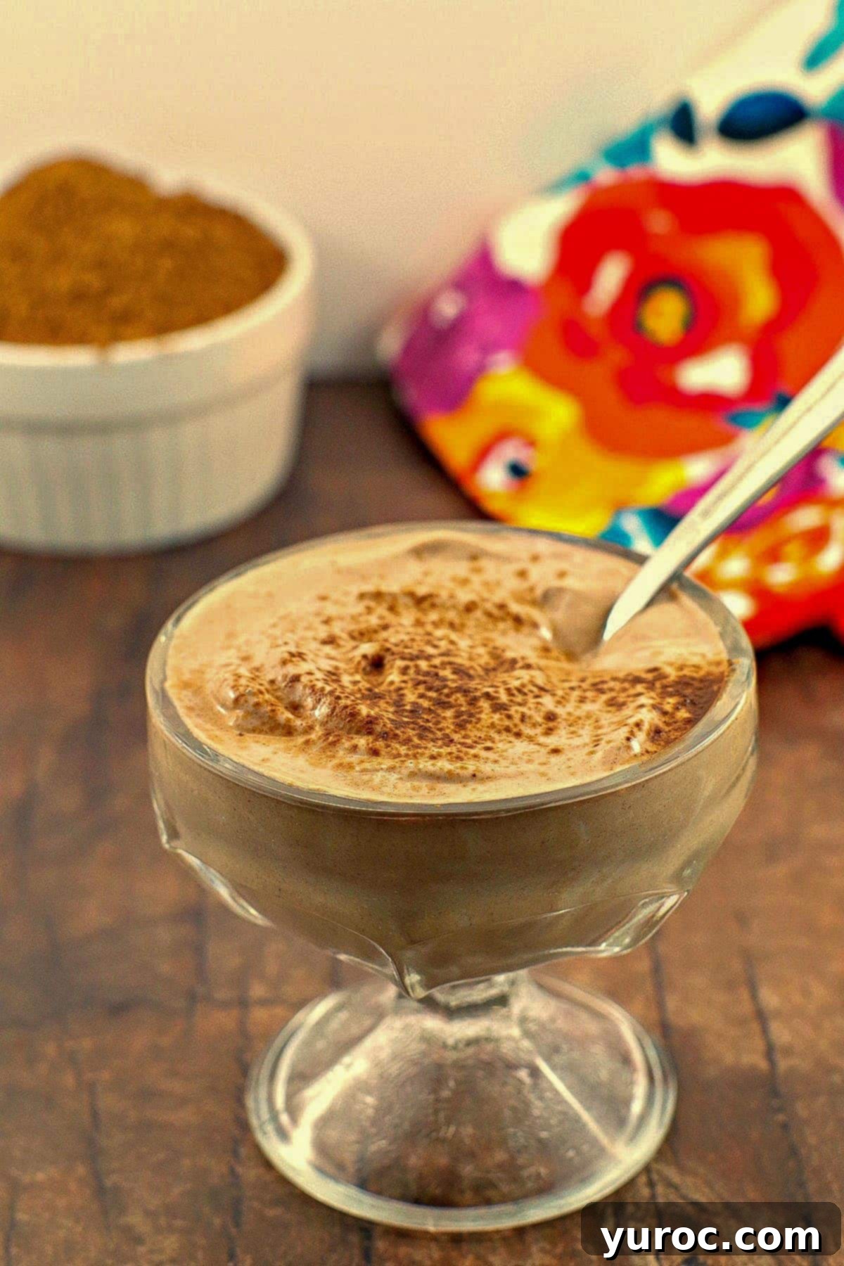 A close-up of creamy chocolate Greek yogurt served in a clear glass dessert bowl, with a spoon resting in it.