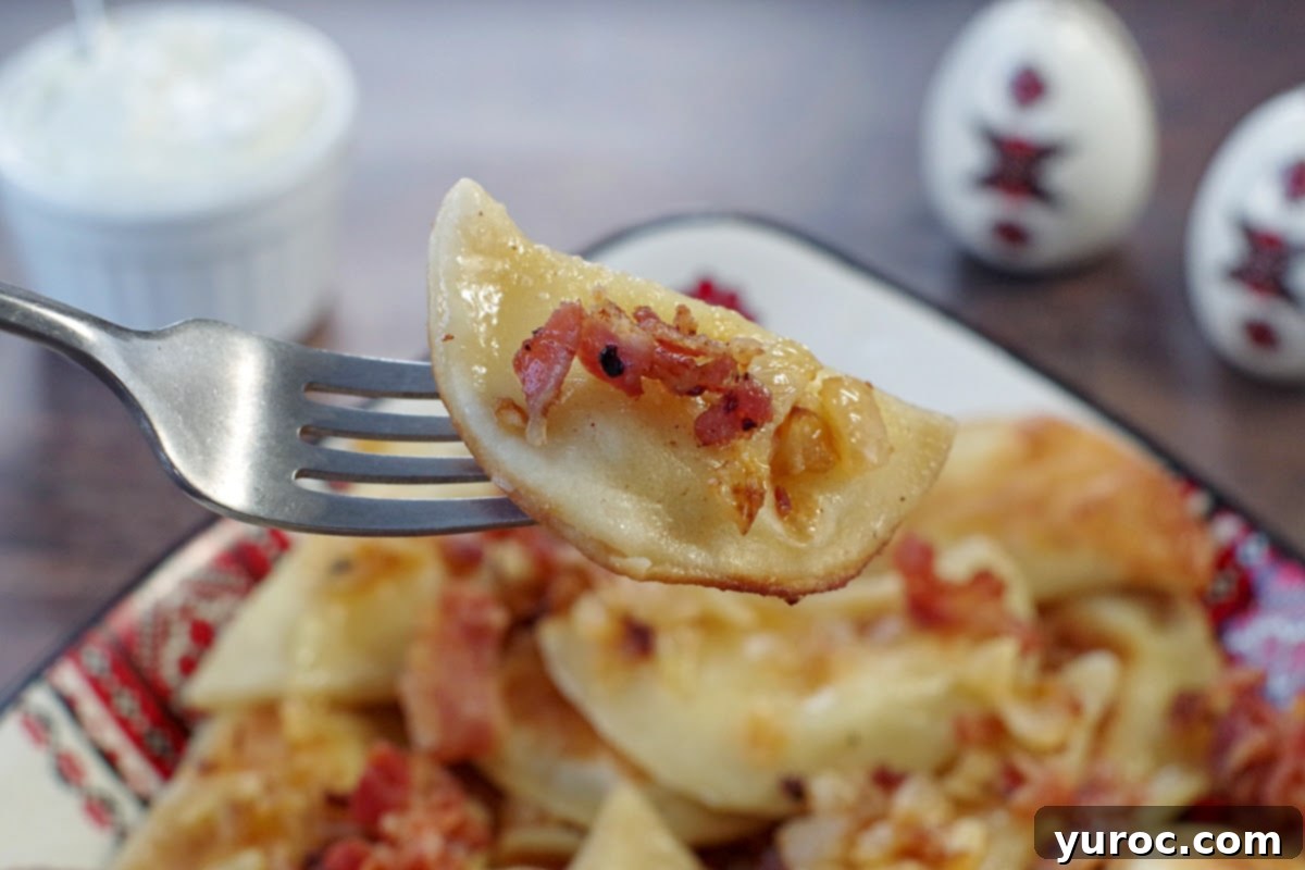 potato and cheese pierogi being held up on a fork over a dish of perogies with bacon and onion with sour cream and ukrainian print salt and pepper shakers in the background