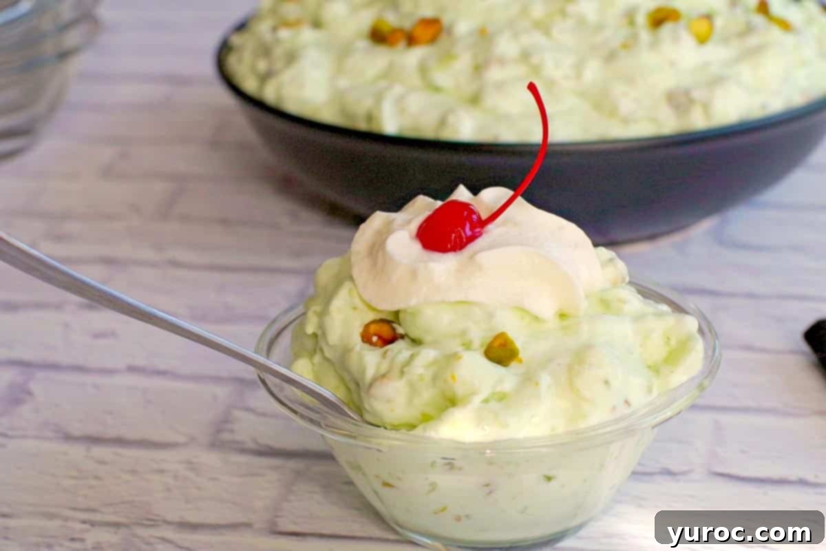 Creamy Pistachio Fluff 2 A serving of Pistachio Watergate salad in a dessert bowl with a spoon, with a larger bowl of pistachio fluff in the background, ready to be served.