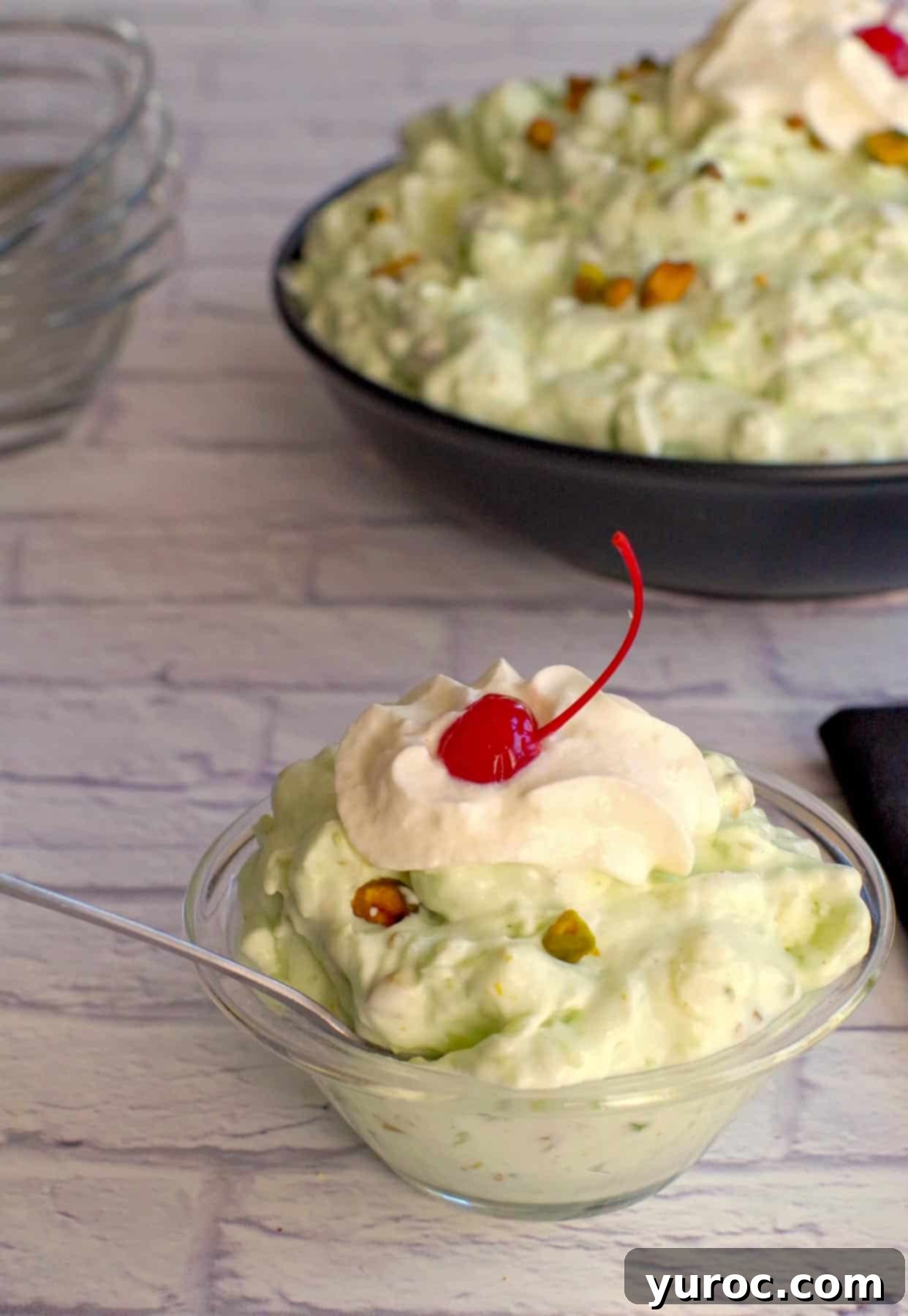 Creamy Pistachio Fluff 11 A close-up shot of Pistachio Watergate salad in a decorative dessert bowl with a spoon, framed by more pistachio fluff in the background, adorned with a maraschino cherry and chopped pistachios.