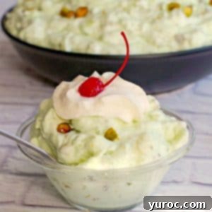 Creamy Pistachio Fluff 12 pistachio fluff in a glass bowl with a larger bowl in the background, garnished with a maraschino cherry and chopped pistachios