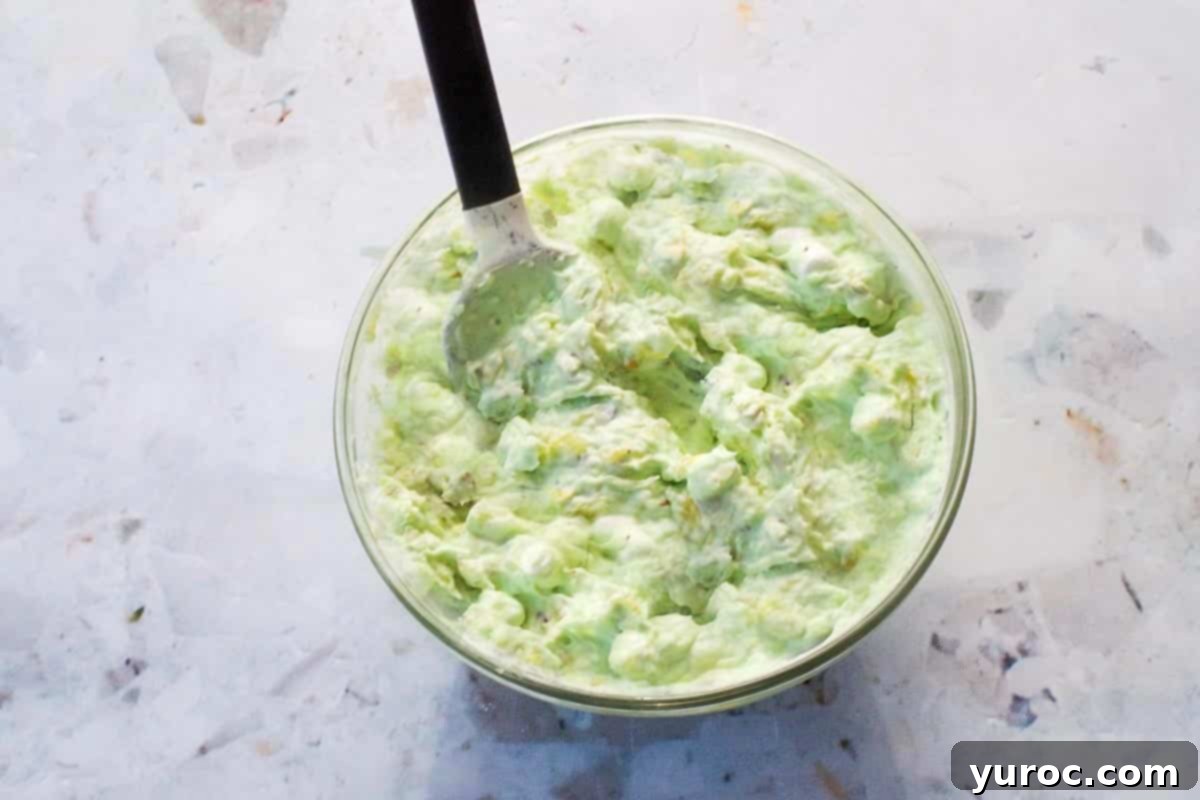 Creamy Pistachio Fluff 8 Finely chopped raw pistachios being stirred into the green fluff mixture in a glass bowl with a spatula, adding texture and more pistachio flavor.