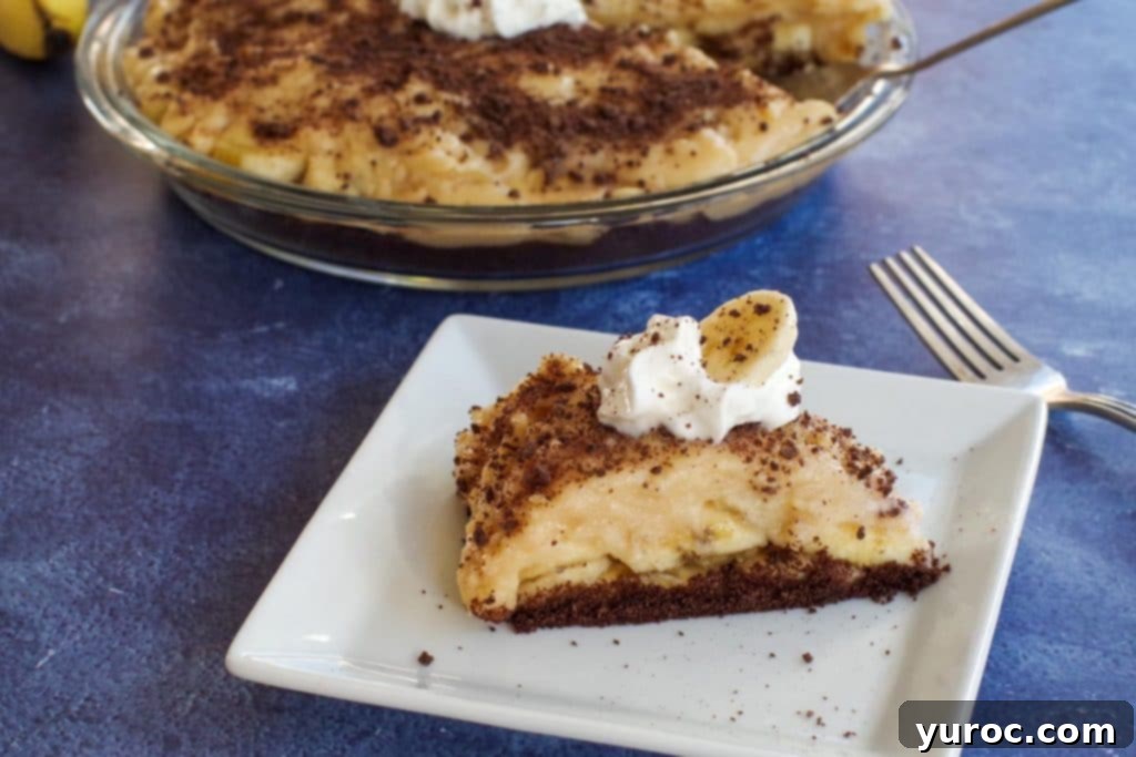 Lightened Up Old Fashioned Banana Cream Pie 1 A perfectly sliced piece of healthy banana cream pie on a white plate, with a whole pie in the soft-focus background.