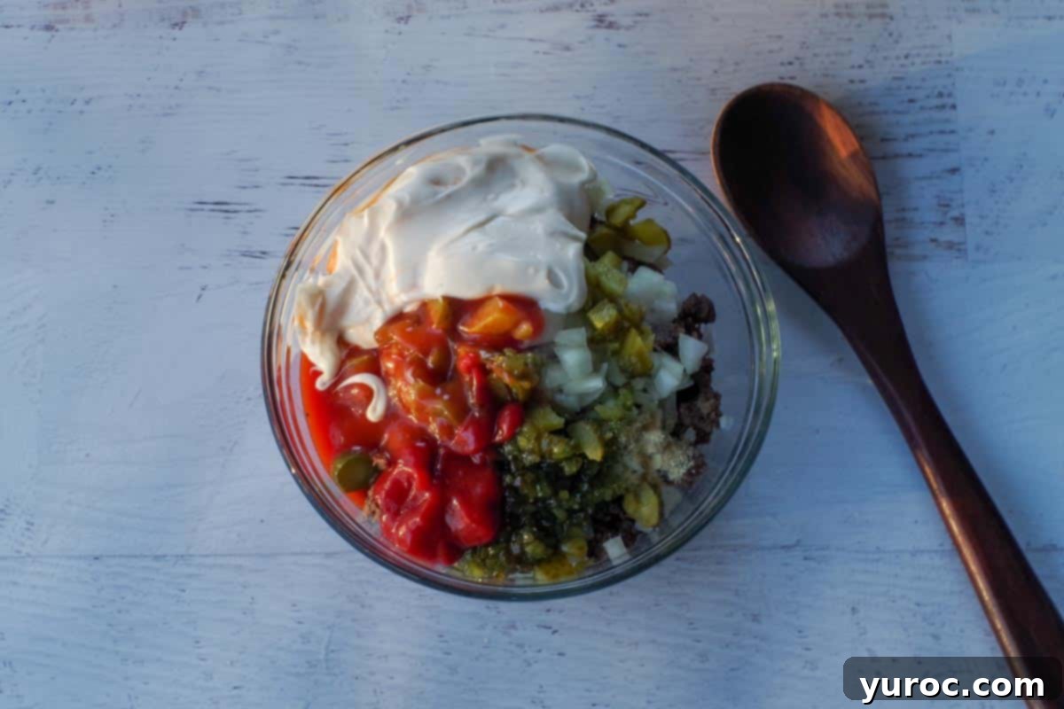 Chopped pickles, onions, and Big Mac dressing (light Catalina salad dressing, mayonnaise, ketchup, relish, onion powder, salt, and vinegar) added to medium glass bowl with cooked ground beef., with a dark brown wooden spoon beside the bowl. 