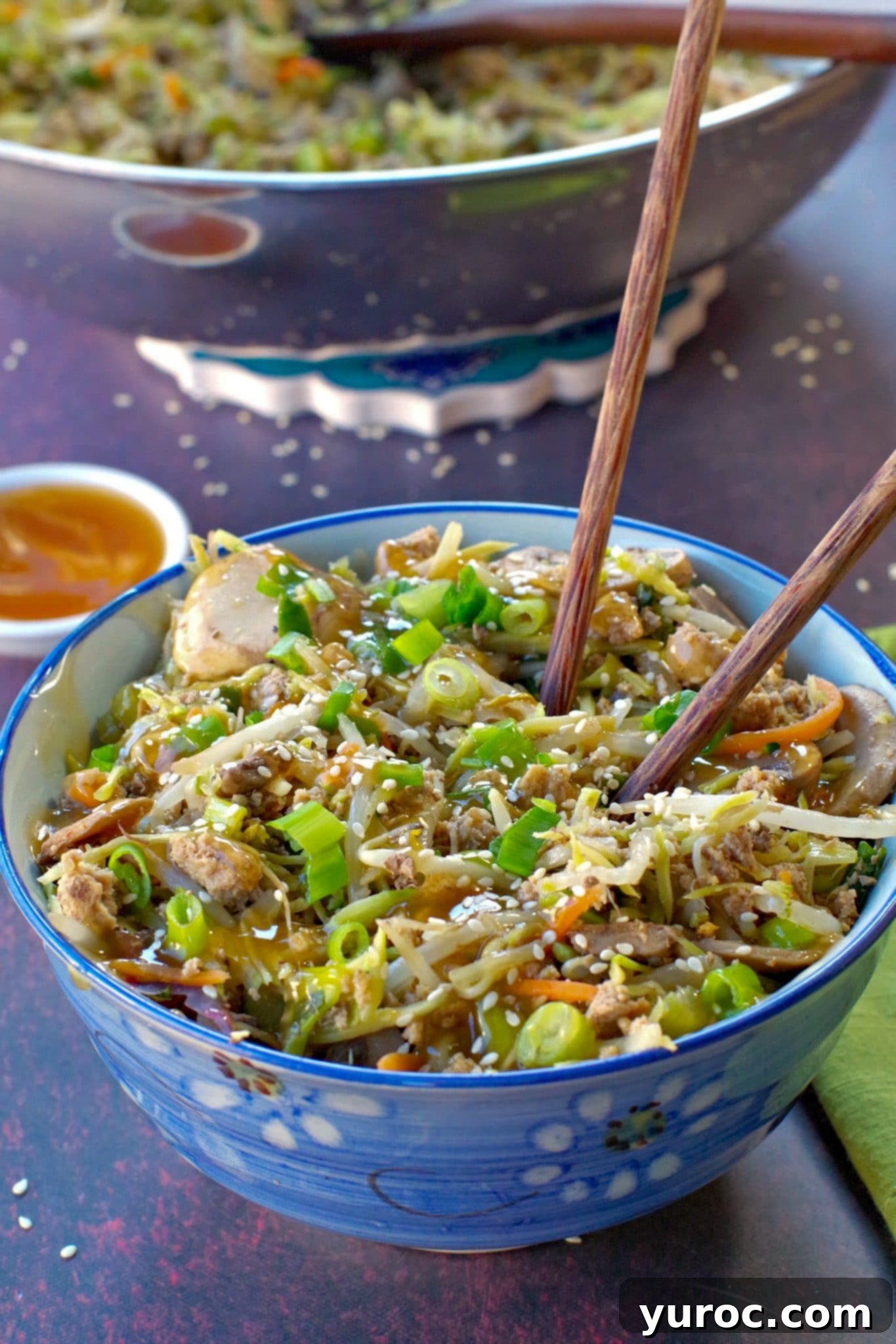 Egg roll in a blue flowered bowl with chopsticks and a small white dish of plum sauce in the background and wok with more egg roll in a bowl in the background