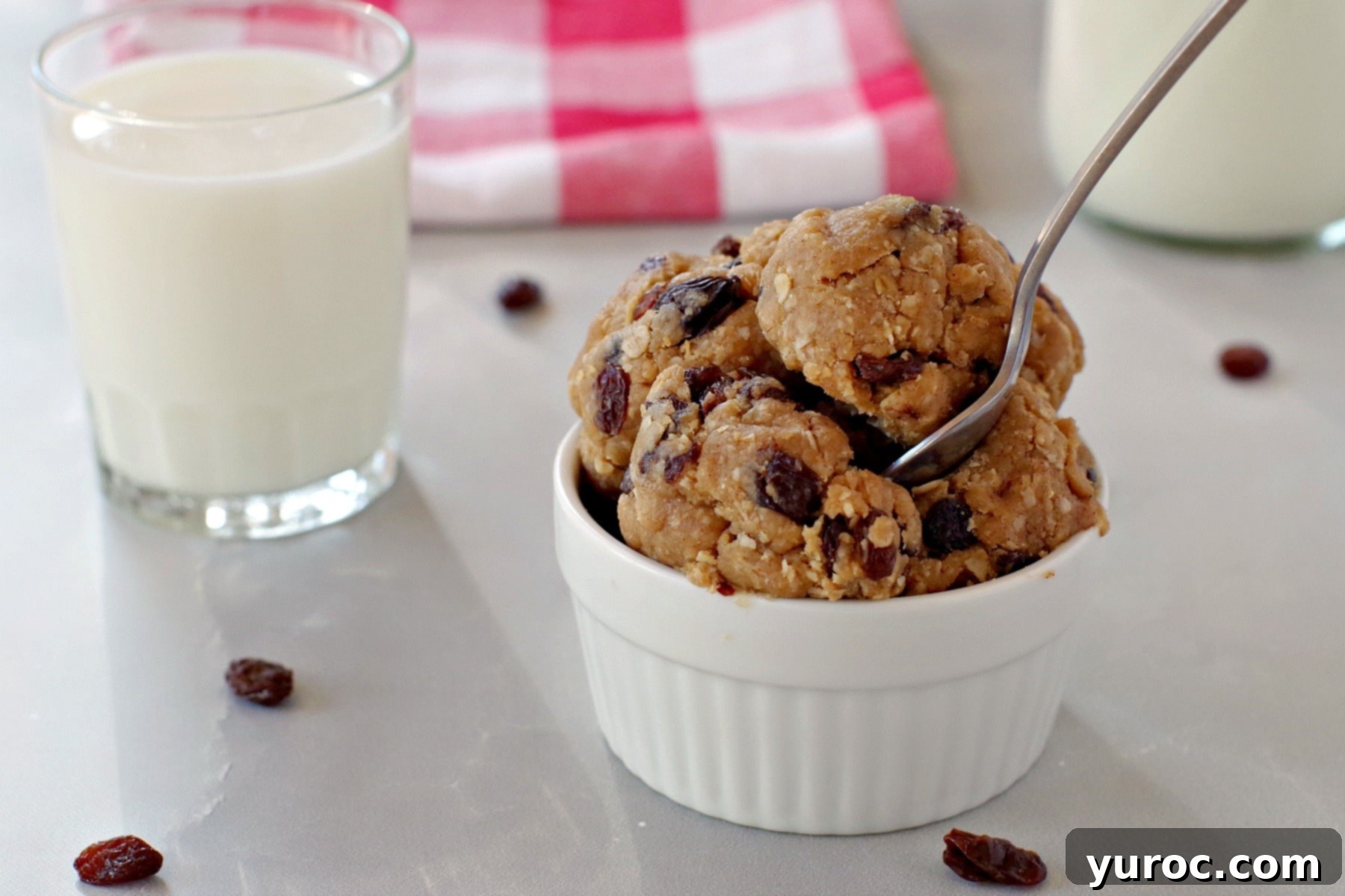 Oatmeal Raisin Dough Delight 2 A white bowl filled with creamy oatmeal raisin edible cookie dough, a spoon resting in it, with a glass of milk in the soft-focus background.
