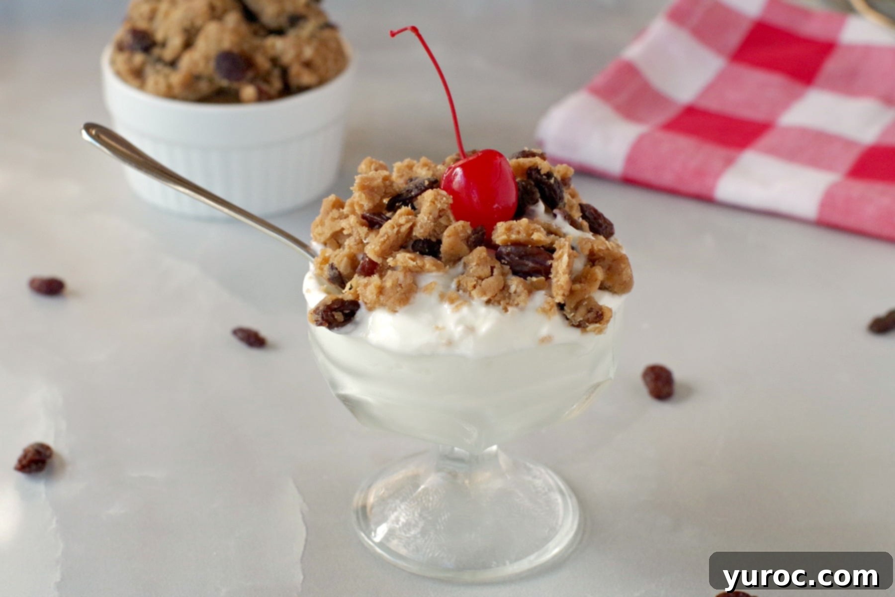 Oatmeal Raisin Dough Delight 5 A glass dish of yogurt topped with crumbled edible cookie dough and a bright red cherry.