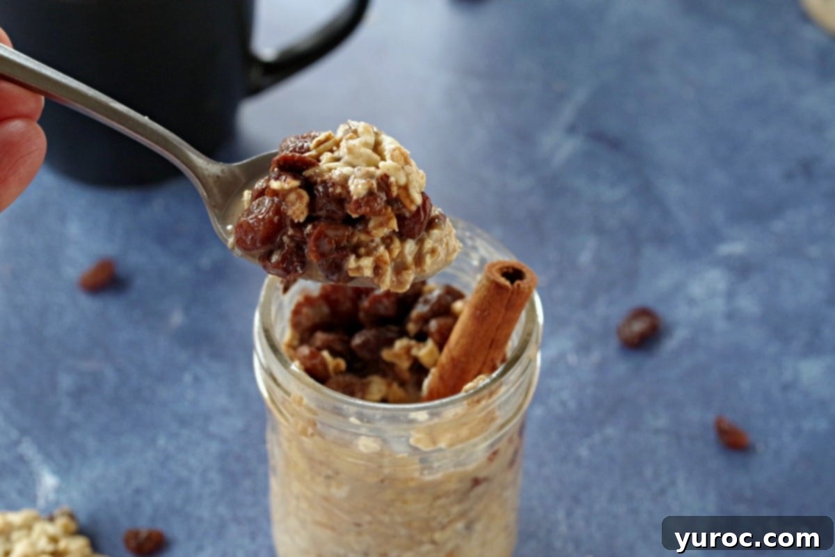 A jar of oatmeal raisin cookie overnight oats with a cinnamon stick, a spoon of oats, and a black coffee mug in the background, all on a blue surface.