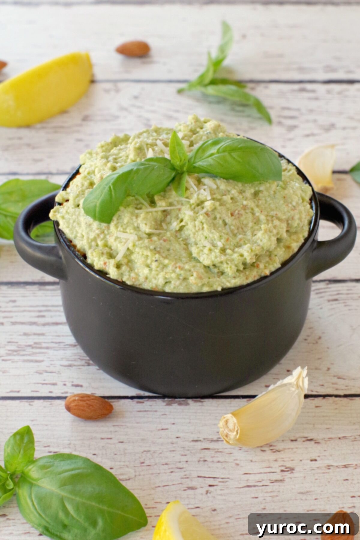 basil pesto with almond in a black dish with almonds, basil, garlic and lemon surrounding the dish