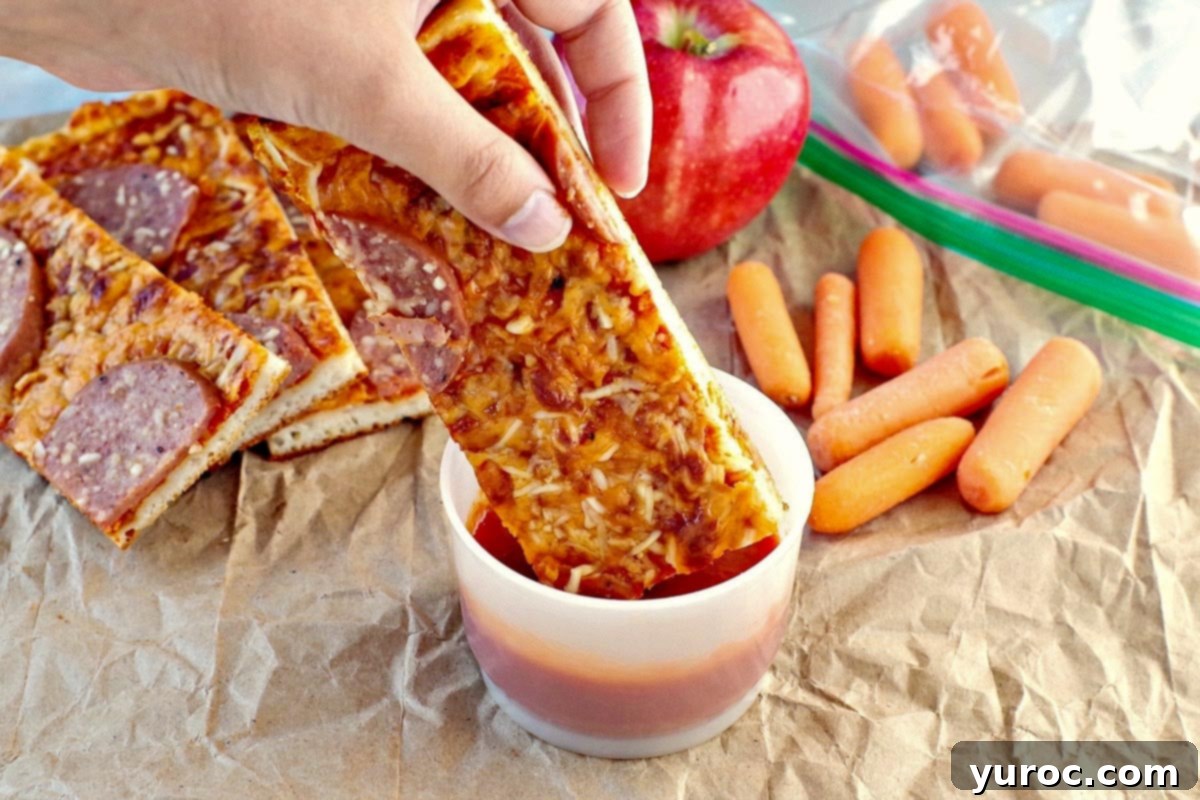 Pizza dipper being dipped into pizza sauce in a small plastic container on a crumpled brown paper bag with and apple and baby carrots in the background.