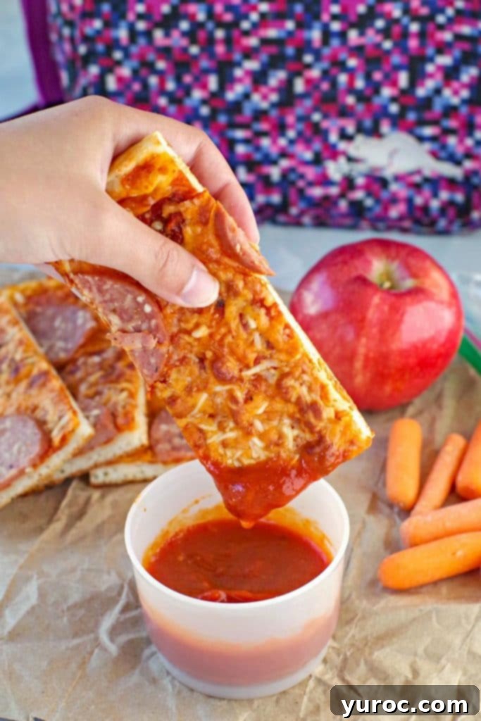 pizza dipper being dipped into pizza sauce with pieces, an apple, carrots and lunch bag in background.