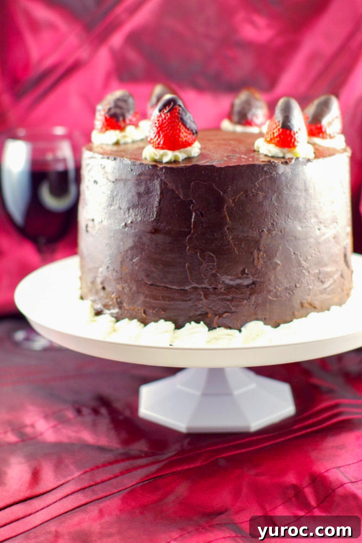 A whole, beautifully garnished Red Wine Chocolate Strawberry cake resting on a white platter, with a glass of rich red wine in the background.