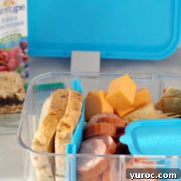 15 No-Prep Healthy School Lunches for Teens 12 Filled Ukrainian Bento box with juice box, date square and bento box lid in background, featuring kubasa, cheddar cheese, dill pickles, rye bread, crackers, mustard and baby carrots