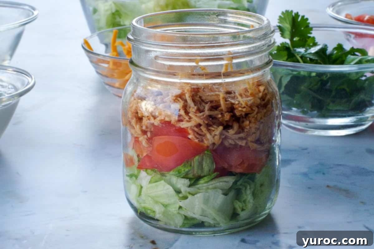 Taco flavored chicken on top of tomatoes and lettuce in glass jar.