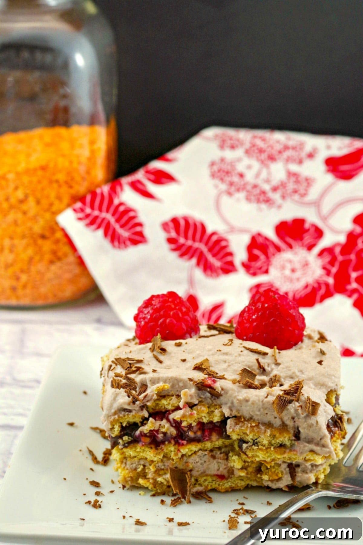 A slice of red lentil chocolate dessert lasagna on a white plate with a jar of red lentils and a red and white tea towel in the background.